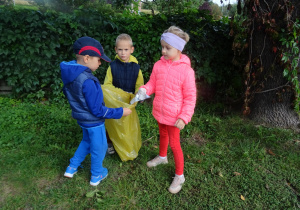 chłopcy trzymają żółty worek na śmieci a dziewczynka przyniosła znalezioną butelkę plastikową z krzaków